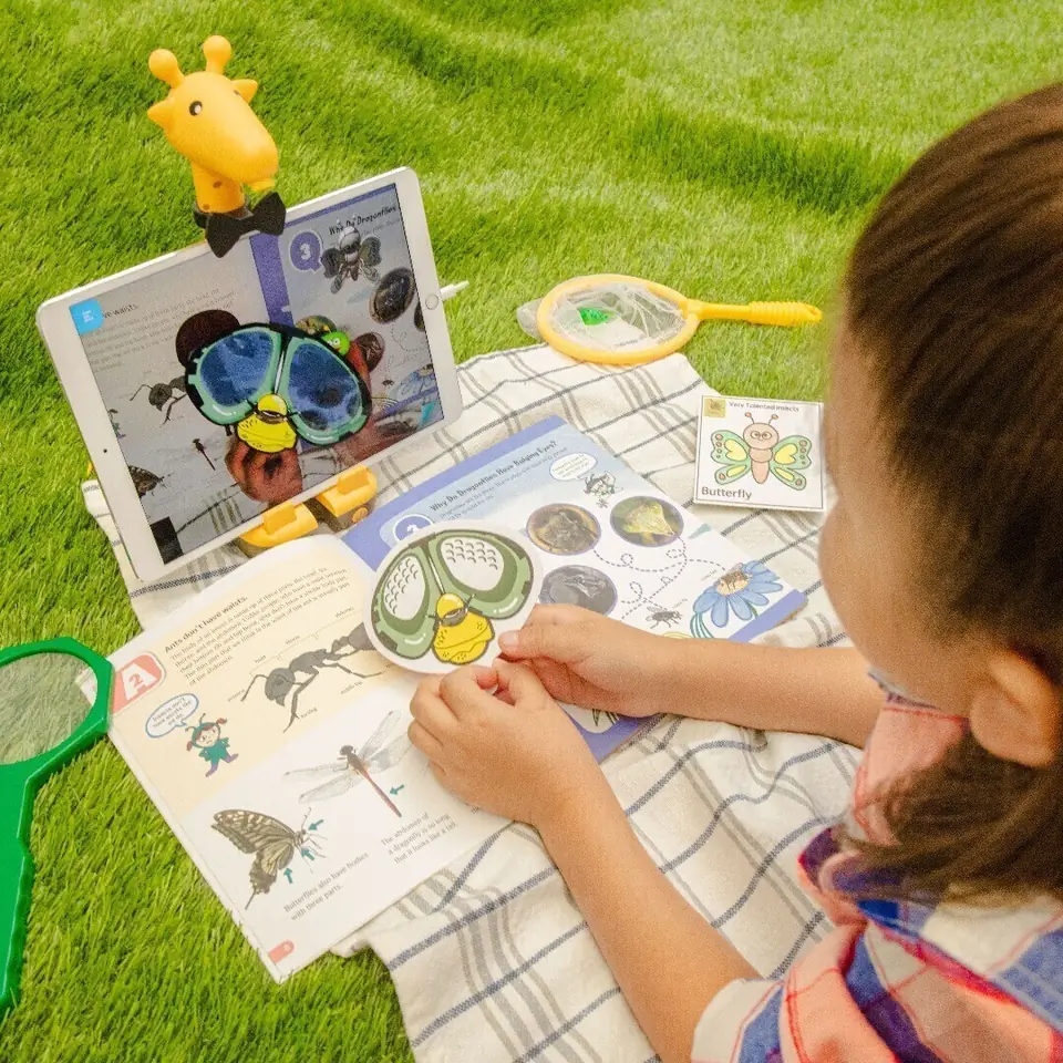 芝生の上で ARpedia を使う子ども。虫の絵本を開き、マーカーを手に持っている。タブレットの画面にARの映像が映っている